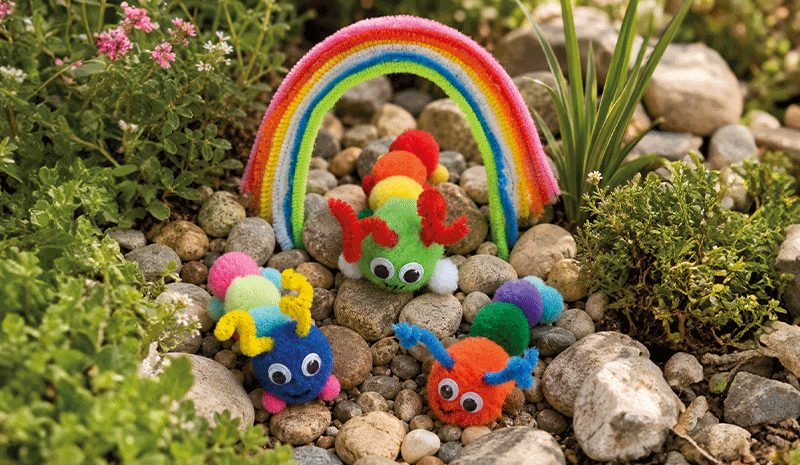 Raupen im Steingarten Bunte Raupen aus Pompons und Pfeifenputzern mit Wackelaugen unter einem Regenbogen in einem Steingarten.