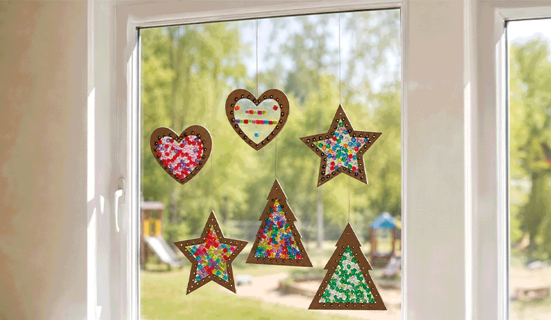 Bunte selbstgebastelte Fensterbilder in Herz-, Stern- und Tannenbaumform mit farbigen Perlen, aufgehängt an einem Fenster.