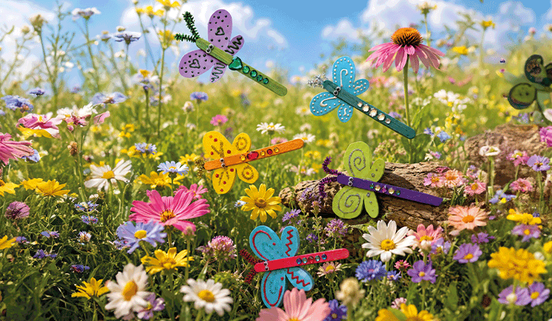 Bunte selbstgebastelte Schmetterlinge aus Bastelstäbchen und dekorierten Flügeln auf einer sommerlichen Blumenwiese.