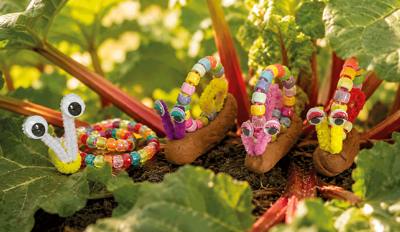 Bunte selbstgebastelte Schnecken aus Perlen, Pfeifenputzern und Wackelaugen zwischen den Blättern einer Rhabarberpflanze.
