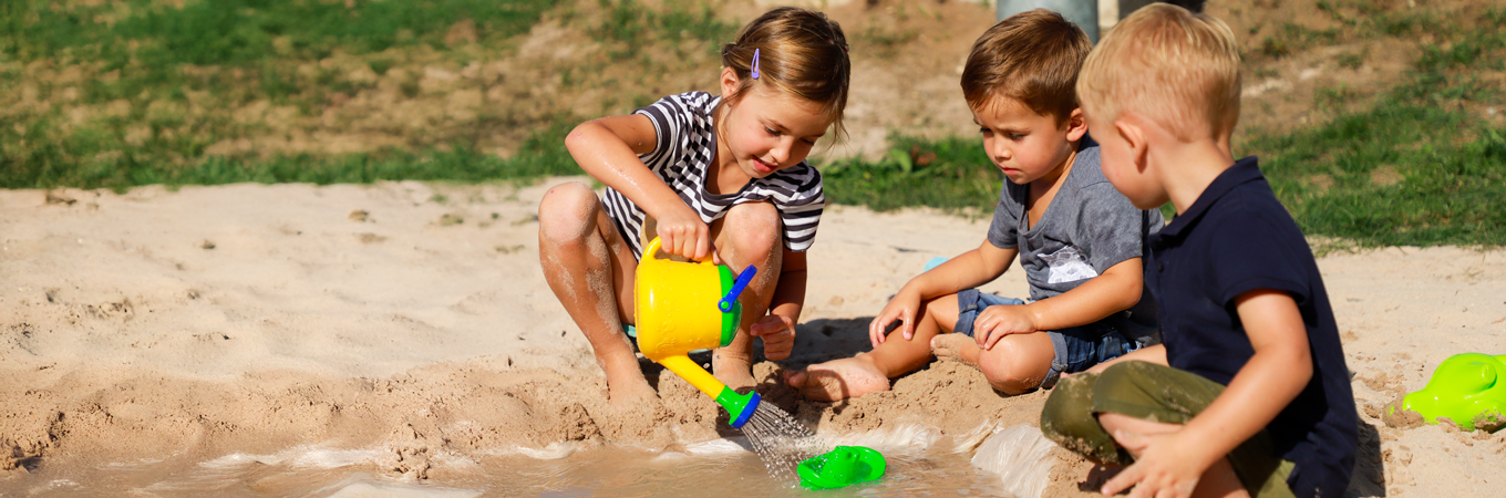Jetzt mit Sand- und Wasserspielmaterial ausstatten Kinder spielen gemeinsam mit Gießkanne und Booten in einer Pfütze im Sand.